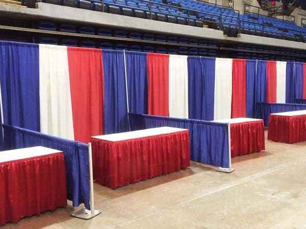 Red, white, and blue pipe and drape backdrop with red-skirted exhibitor tables in an arena